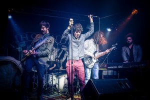 Drei Männer auf einer Bühne im Spotlight, rundum dunkel, Mikrophone, E-Gitarren und Lautsprecher